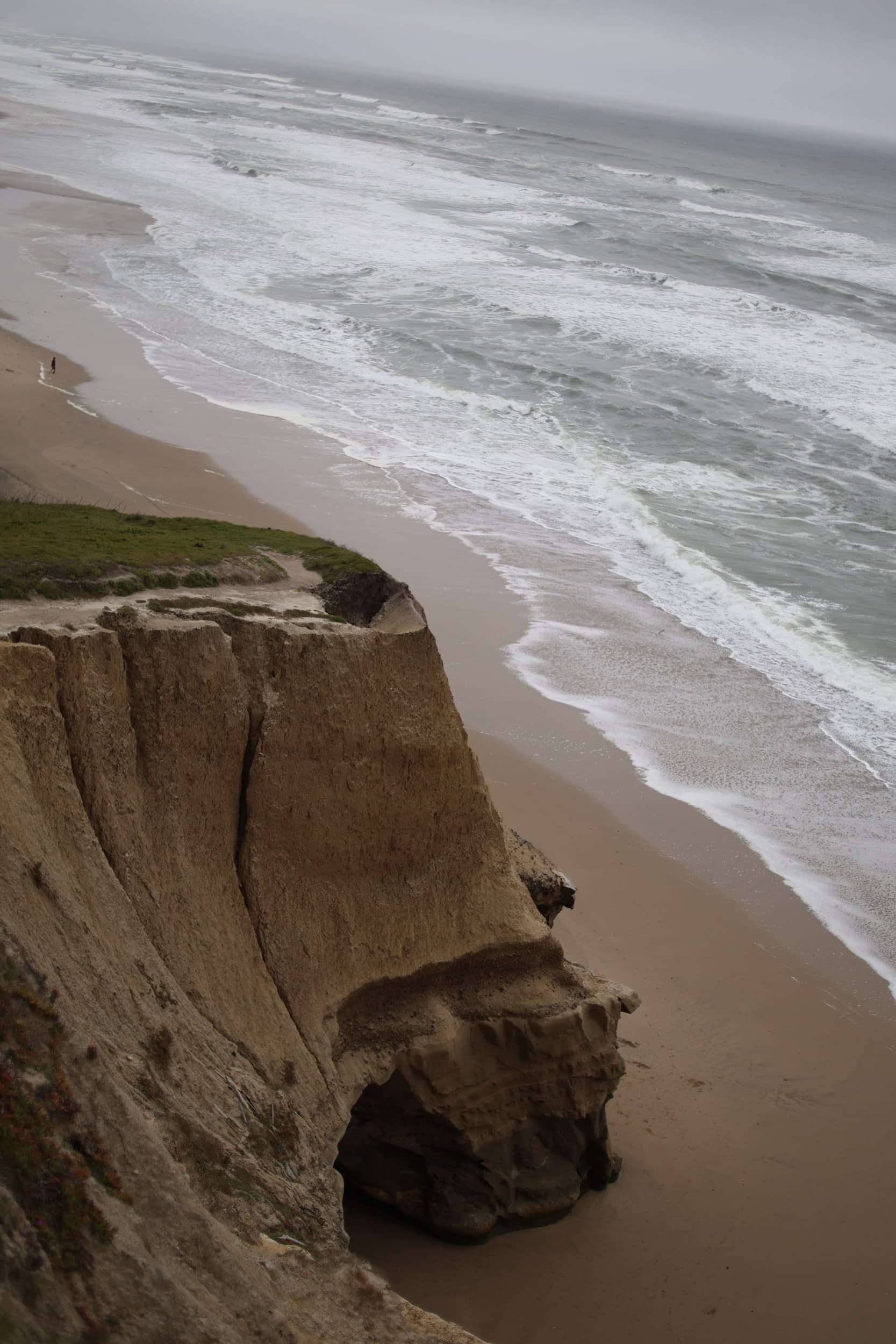 Waves rolling onto a beach below a tall sandstone cliff on an overcast day