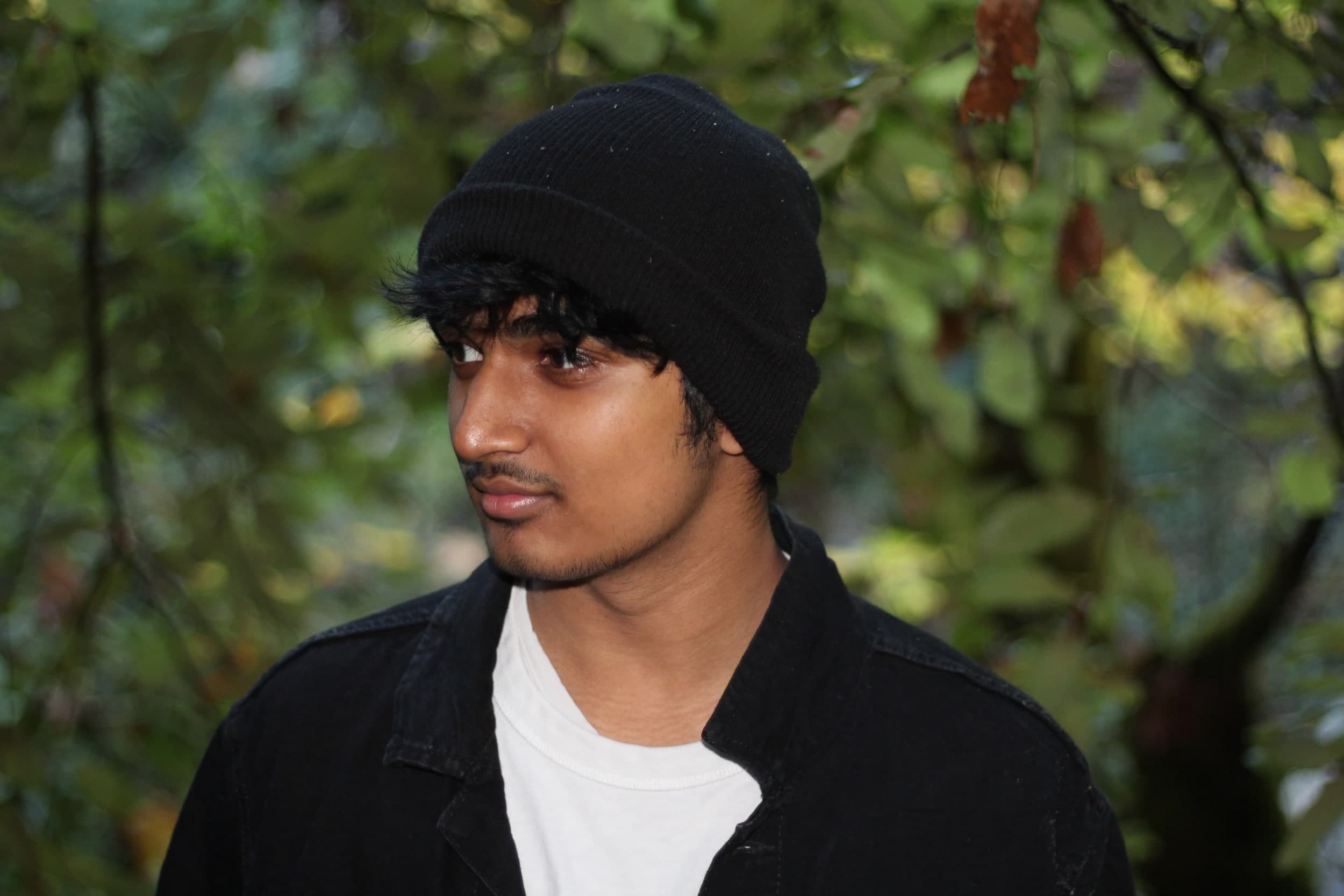 Portrait of a person in a black beanie and jacket looking to the side, with leafy trees behind