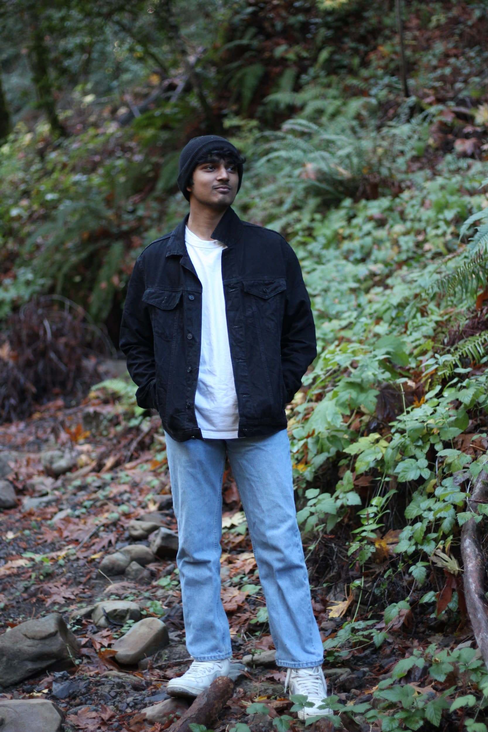 A person in a black denim jacket and light jeans standing on a forest trail