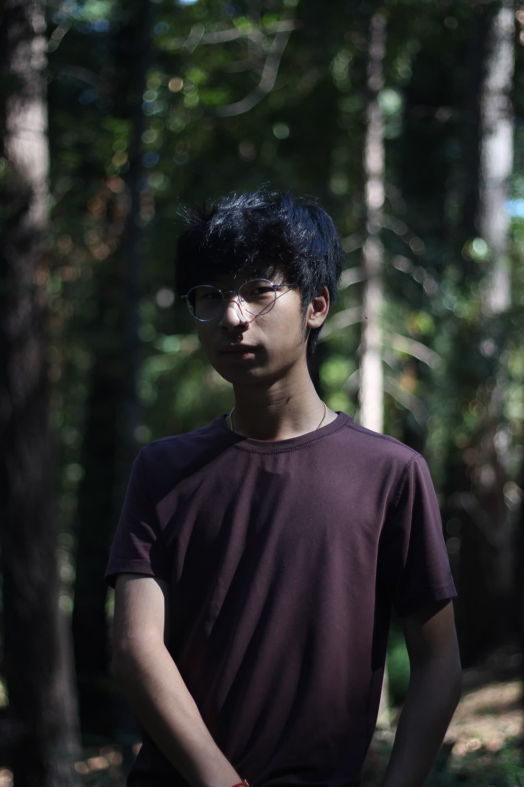 Portrait of a person in round glasses and a maroon T-shirt standing in a sunlit forest