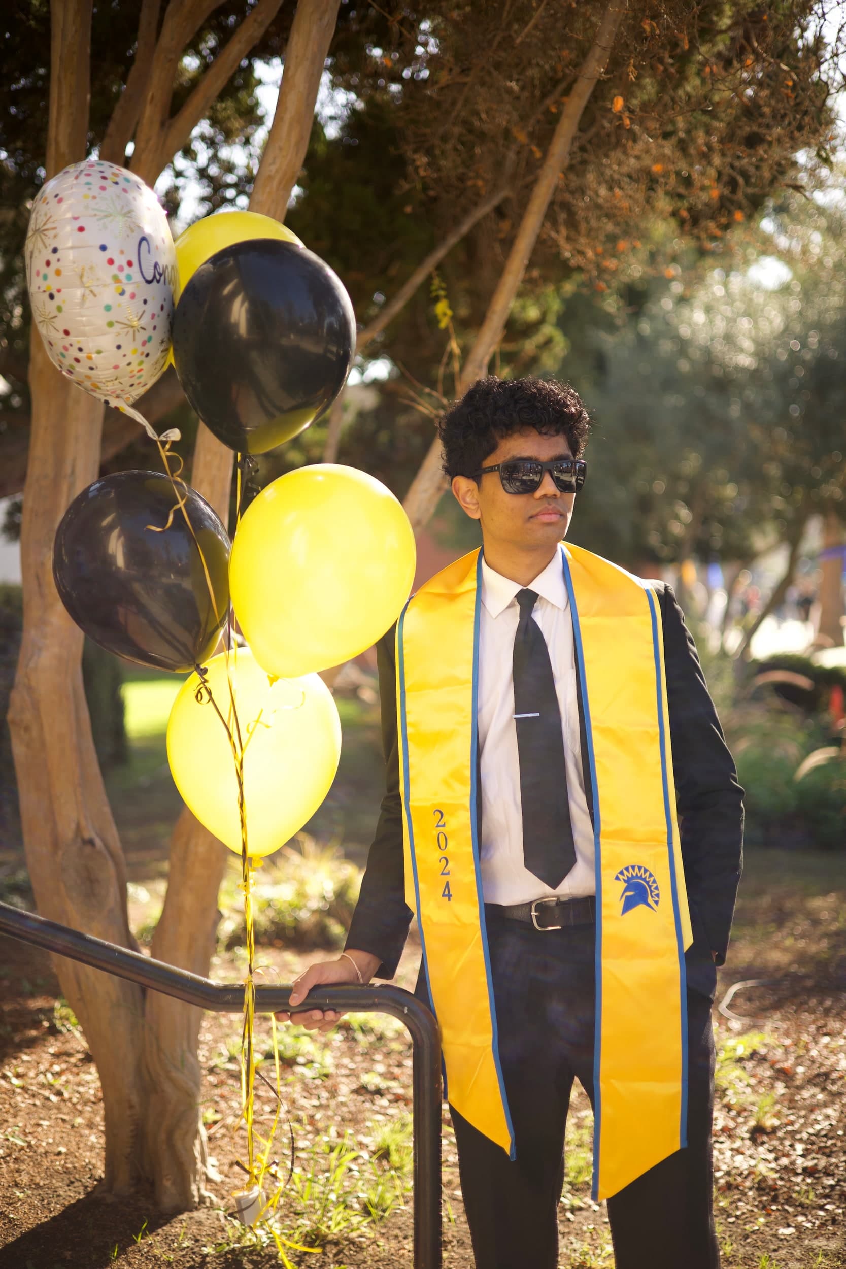 A person in a suit, sunglasses and a gold 2024 graduation stole standing beside yellow and black balloons