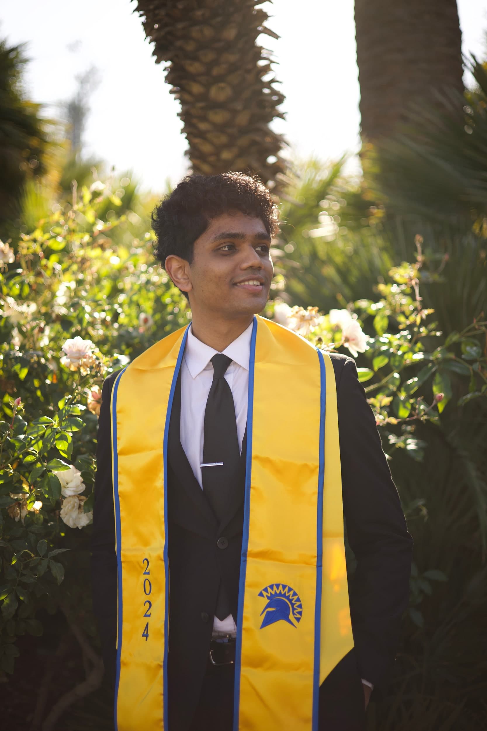 A smiling person in a suit and gold 2024 graduation stole in front of white roses and palm trees