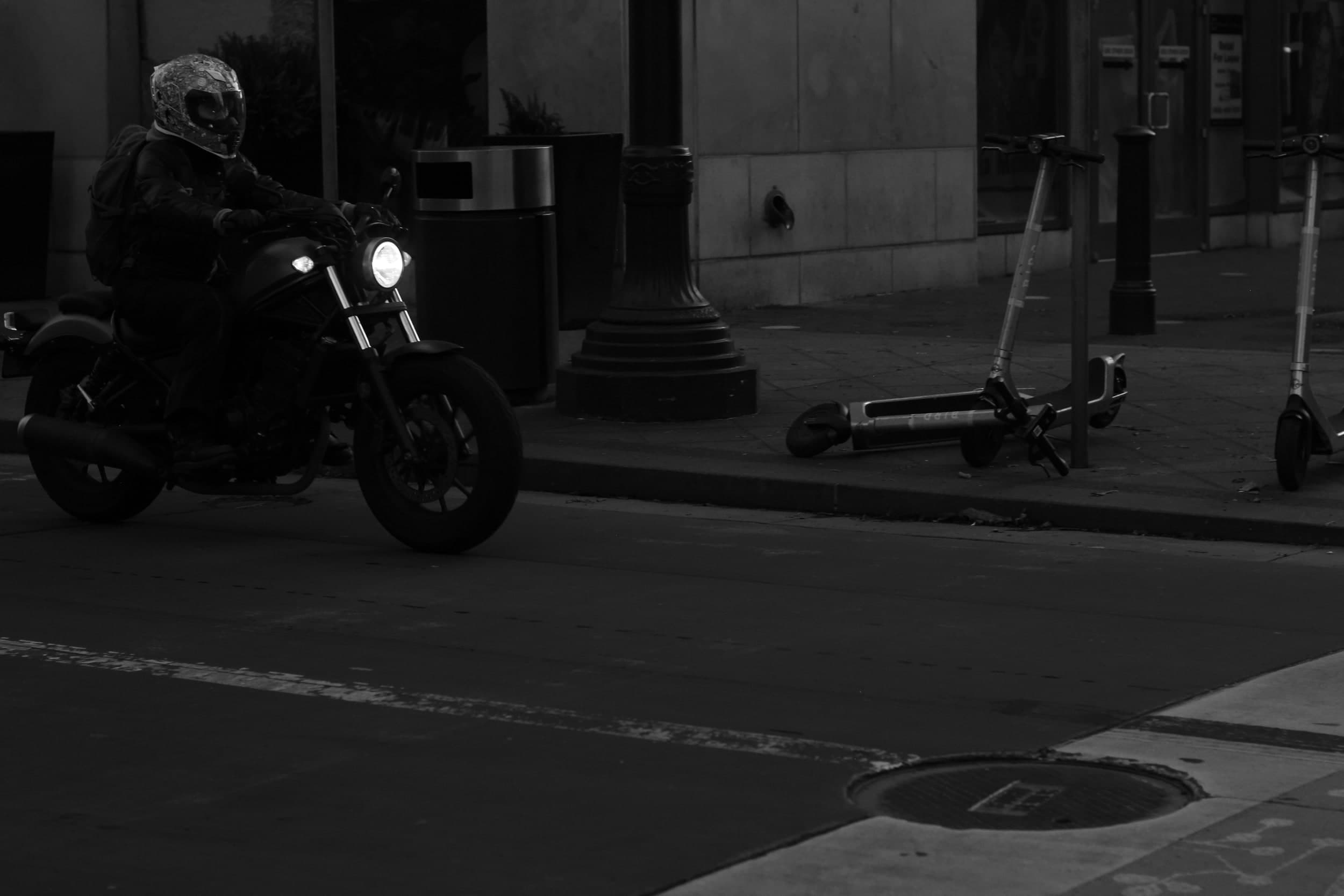 Black-and-white photo of a motorcyclist with a bright headlight riding past parked scooters
