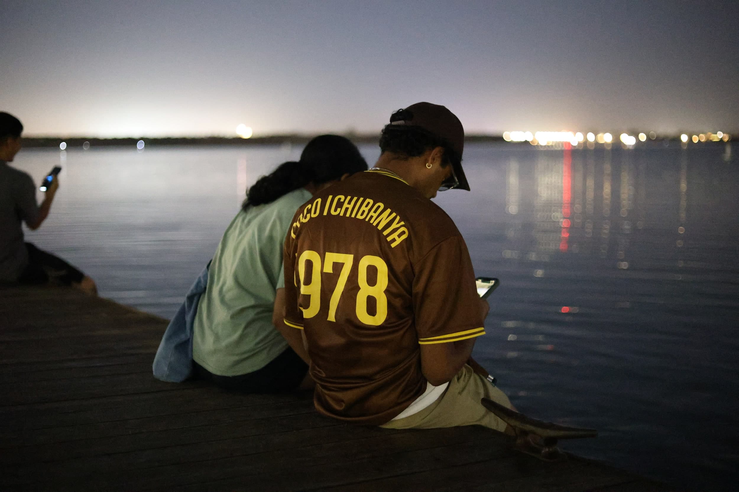 Two people sitting on the edge of a pier at night looking at their phones, with lights across the water
