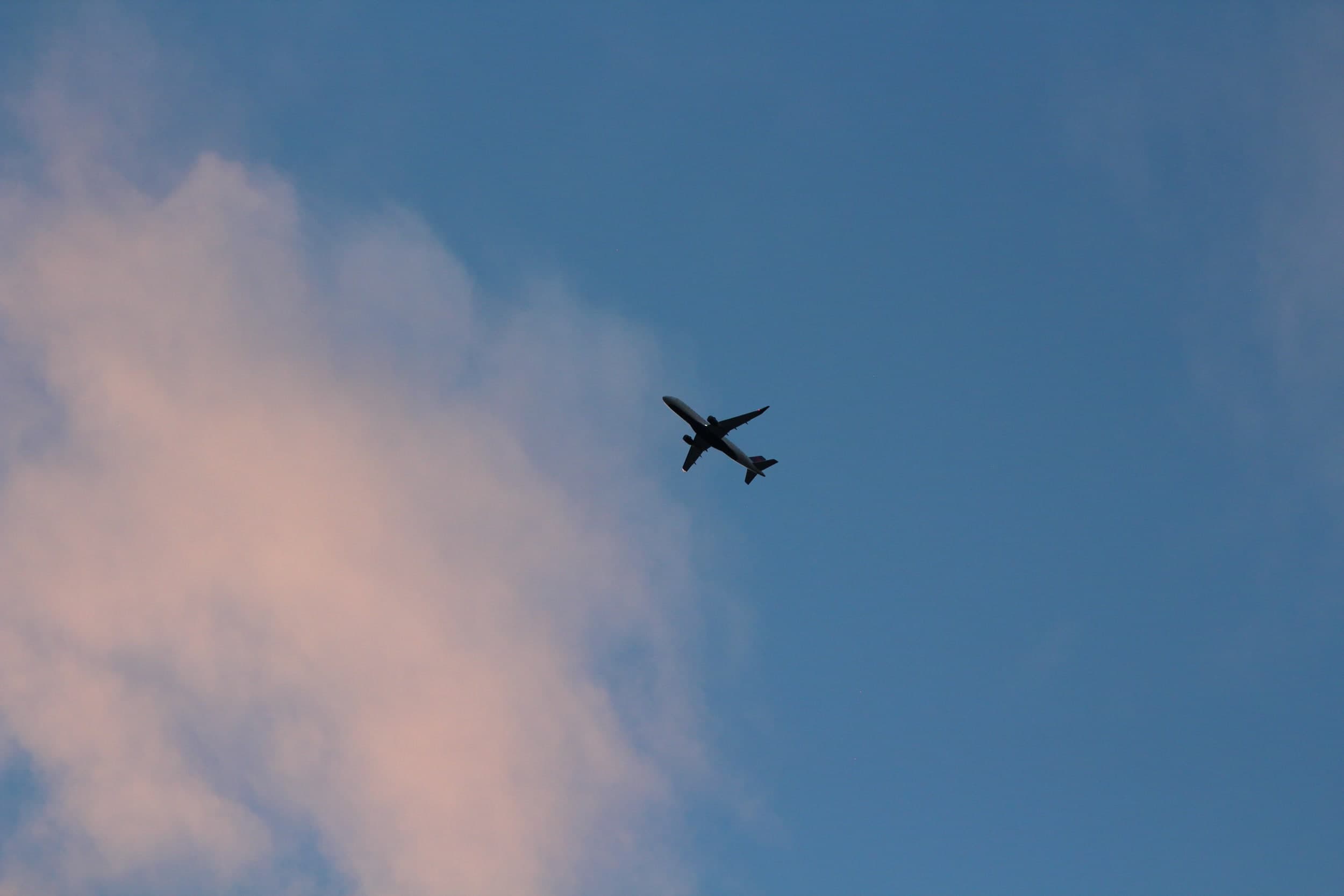A plane flying overhead against a blue sky beside a pink-tinged cloud