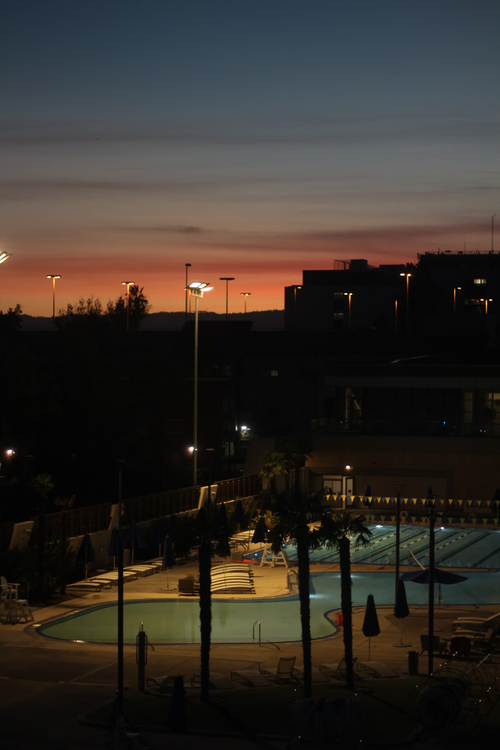 An outdoor swimming pool lit at dusk, with palm trees and a red sunset behind the buildings