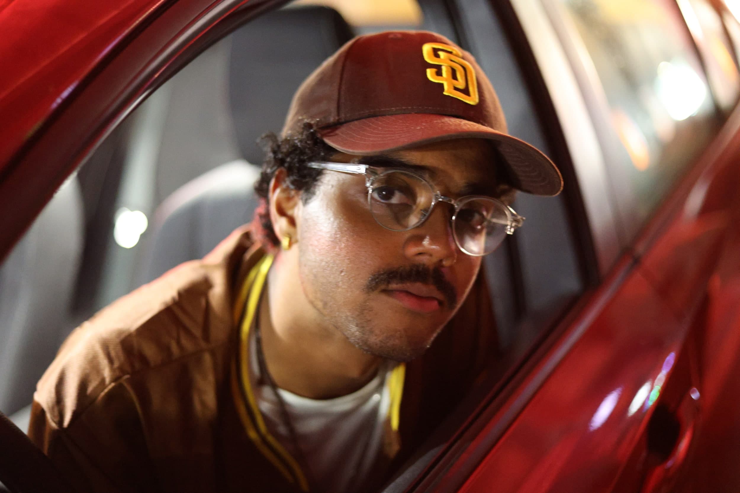 Close-up of a person in a San Diego Padres cap and clear glasses looking out of a red car window