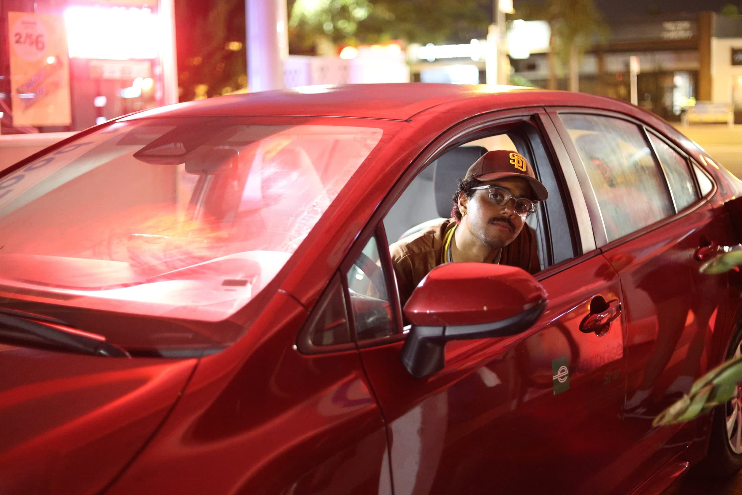 A person in a San Diego Padres cap and clear glasses looking out of the driver's window of a red car at night