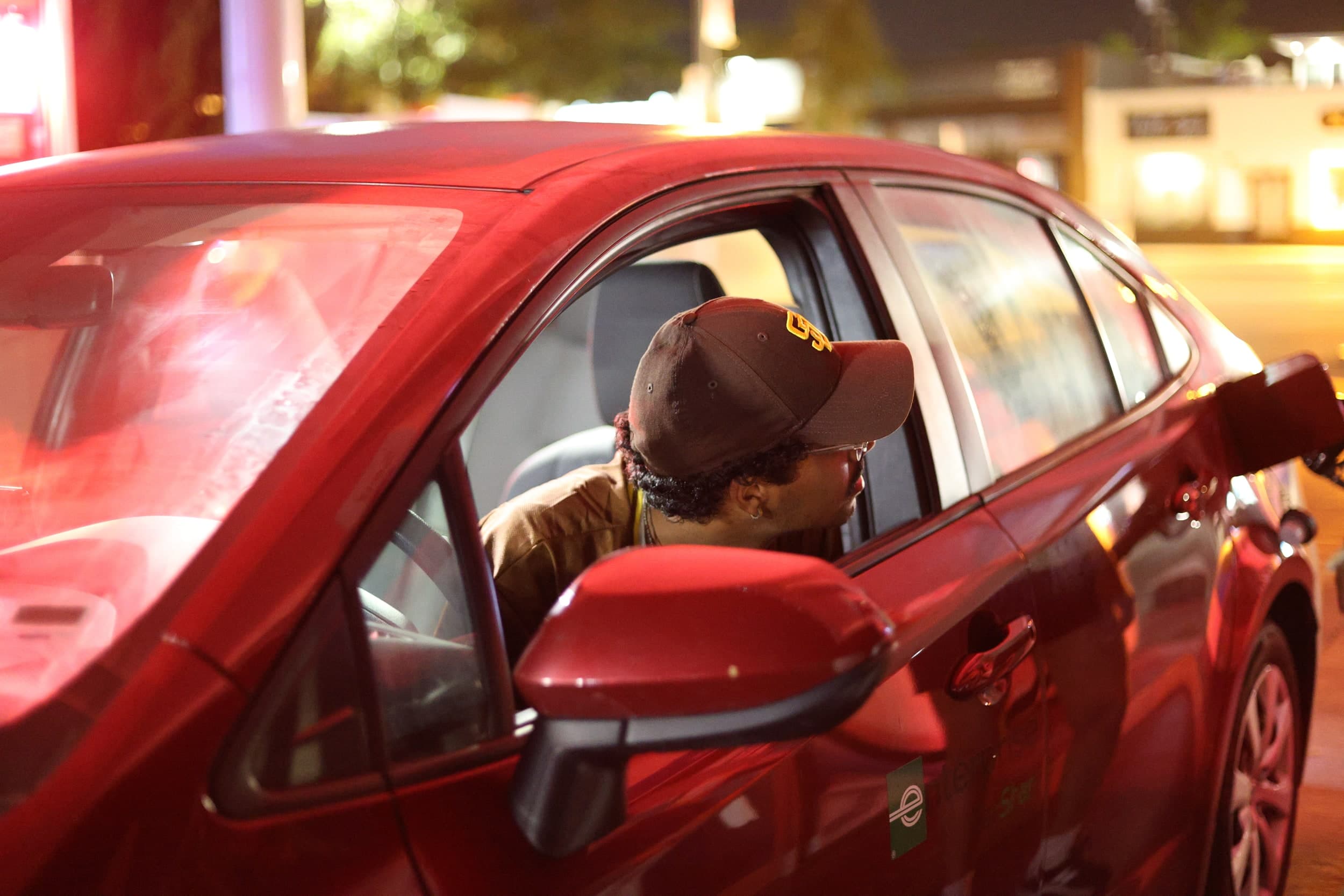 A person in a brown cap leaning out of the window of a red car at night