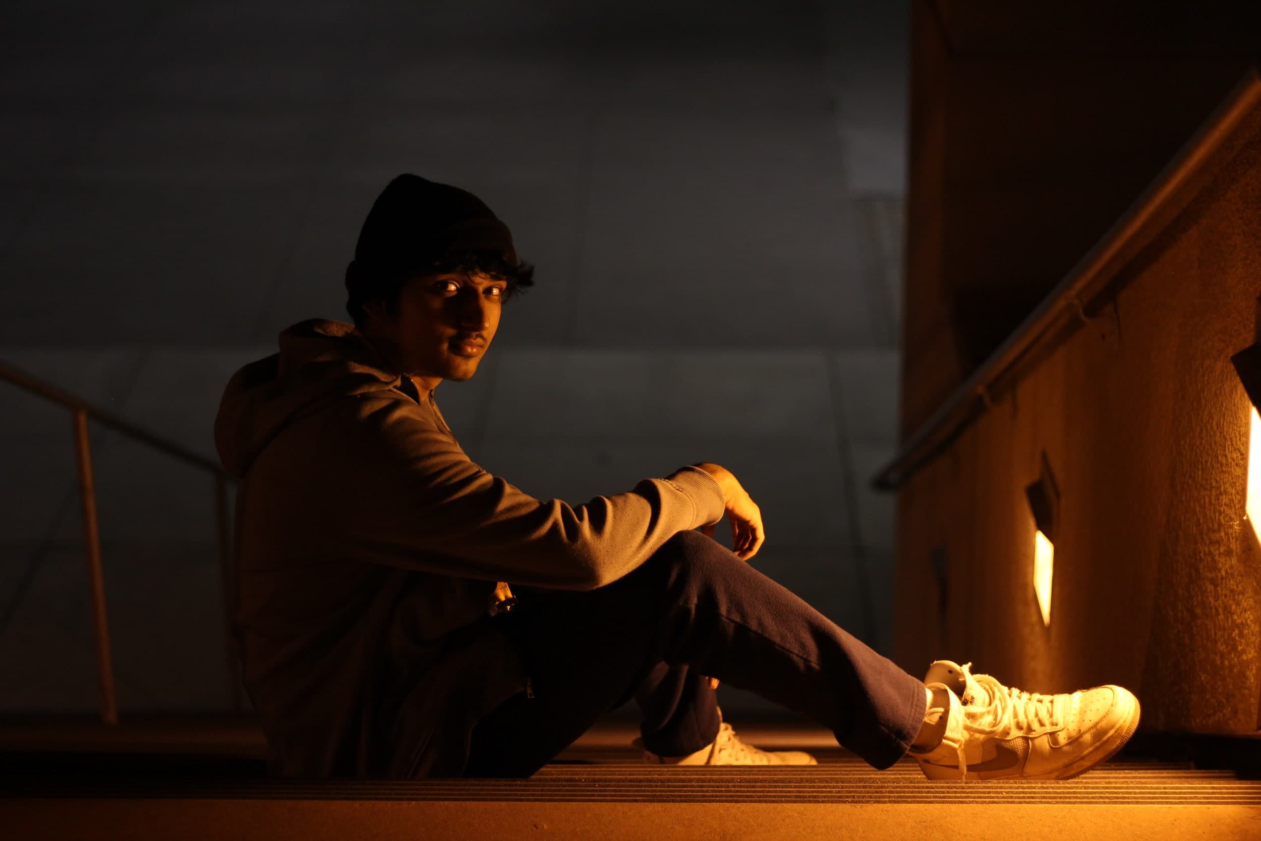 A person in a beanie and hoodie sitting on stairs in warm amber light at night