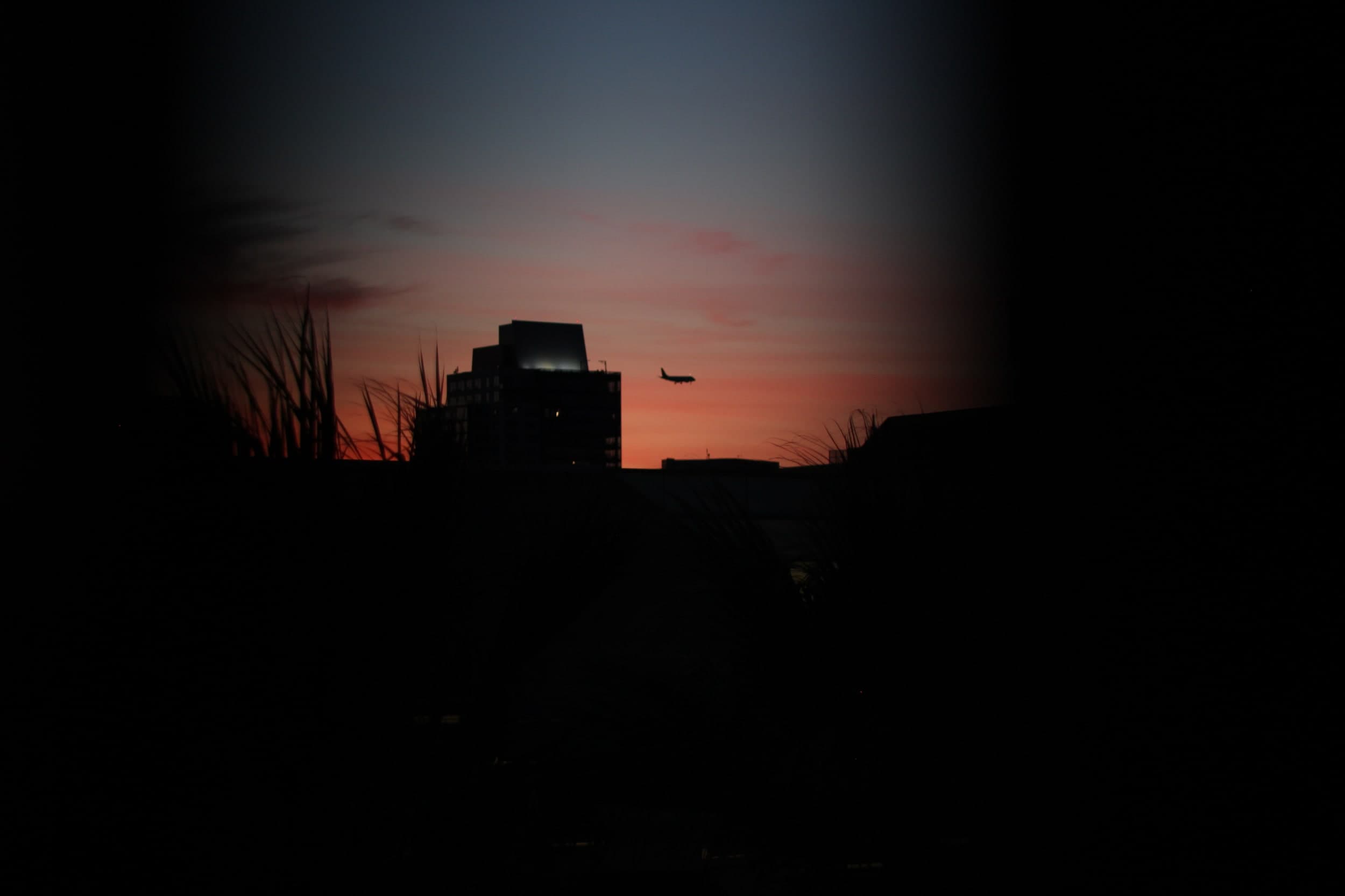 A plane descending past a building silhouetted against a red sunset