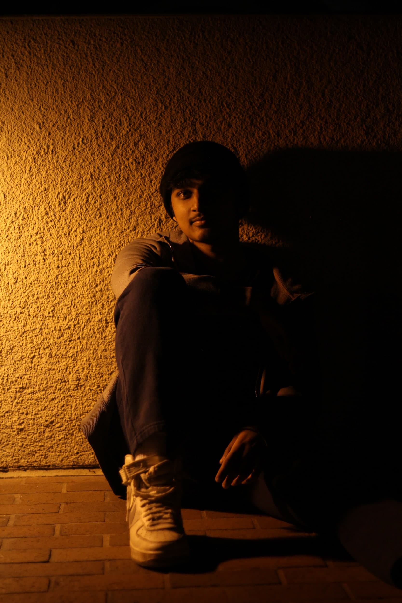 A person in a beanie sitting against a textured wall lit in amber, half in shadow